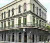 The image is a colorized vintage photo of a two-story corner building with a wrought-iron balcony characteristic of an old urban neighborhood