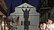 The image shows a striking shadow of a human figure with raised arms projected onto the facade of a white building with a clock tower at dusk.
