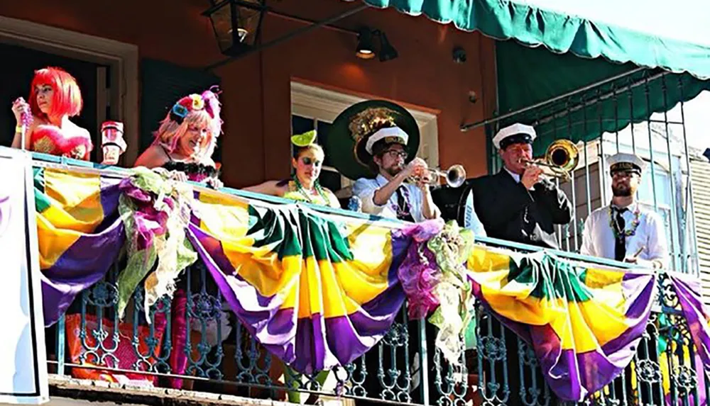 A group of colorfully dressed people and musicians celebrate on a balcony adorned with Mardi Gras colors of purple green and gold