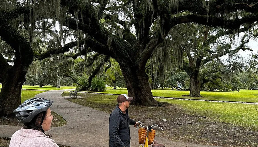 Two people with bicycles are pausing to admire an expansive park with Spanish moss-draped live oak trees.