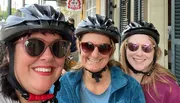 Three smiling individuals wearing bike helmets and sunglasses are posing for a selfie on a city street.