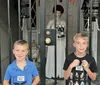 Two smiling boys stand in front of an eerie gated doorway where a creepy doll-like figure holding a phone is flanked by two hanging birdcages