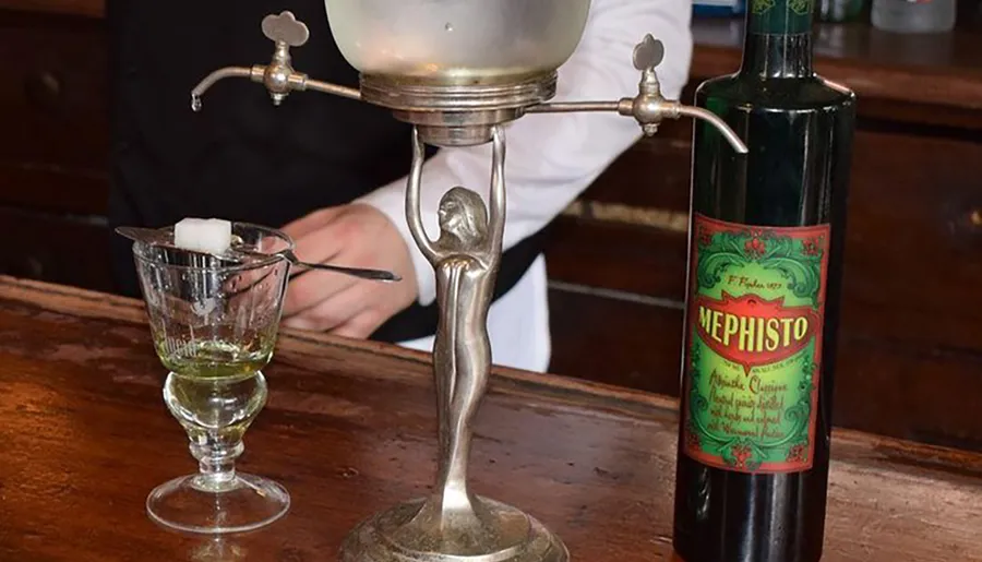 A traditional absinthe fountain is dripping water over a sugar cube on a slotted spoon into a glass of absinthe, with a bottle of Mephisto absinthe next to it.