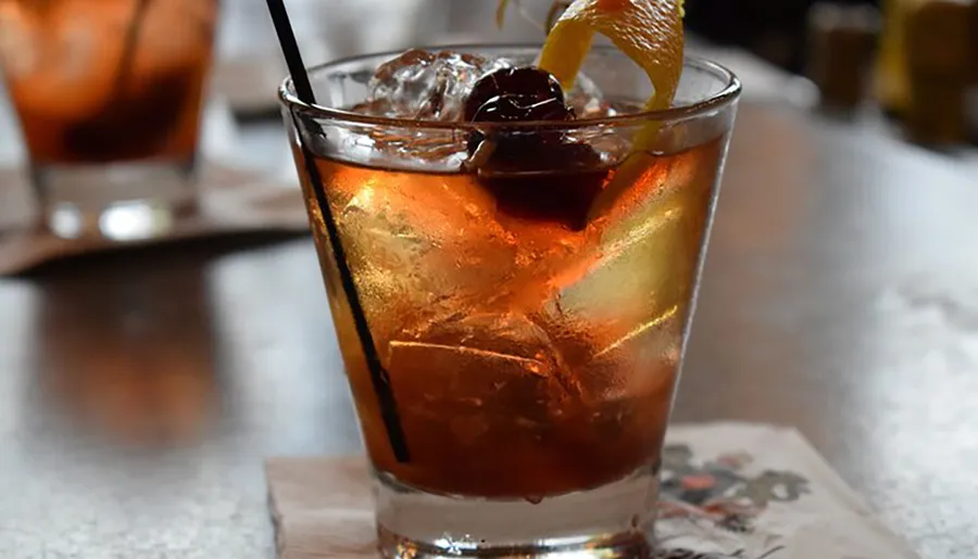 The image shows a close-up of a chilled cocktail with ice cubes, a cherry, and an orange peel garnish, served in a clear glass with a black straw.