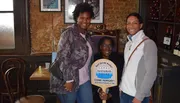 Three smiling people pose inside a rustic-looking establishment, with one holding a sign that reads 