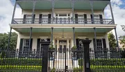 This image features a two-story house with a green-tinted roof and ornate black wrought-iron balcony railings, set behind a decorative black iron fence, exuding classic Southern charm.