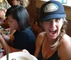 A woman in a hat is making an excited face at the camera while sitting at a table with others who are focused on their phones and drinks