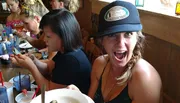 A woman in a hat is making an excited face at the camera while sitting at a table with others who are focused on their phones and drinks.