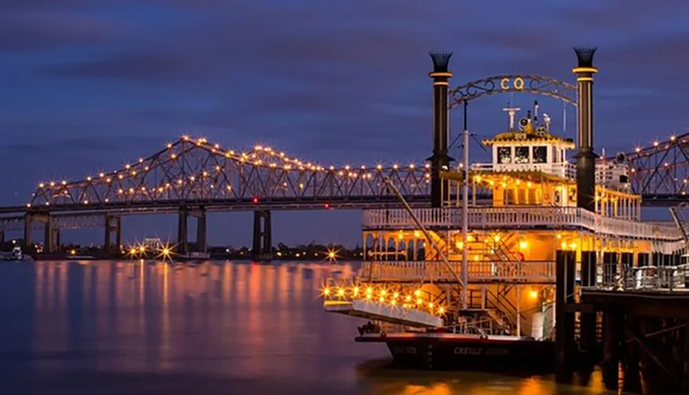 A paddlewheel riverboat is moored at a dock in the evening illuminated by lights with a lit-up truss bridge in the background