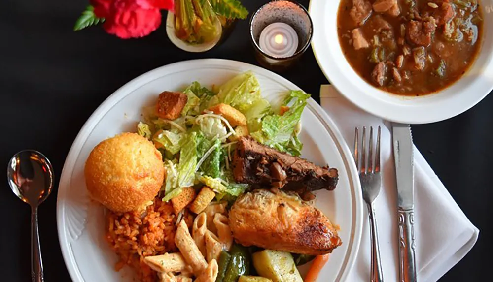 This image shows a plate of diverse foods including salad rice grilled chicken vegetables a roll and a side of stew served with utensils and a lit candle on the table for ambiance