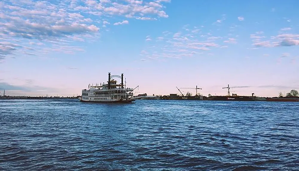 A classic paddle steamer sails on a river against a backdrop of scattered clouds and construction cranes on the distant shore