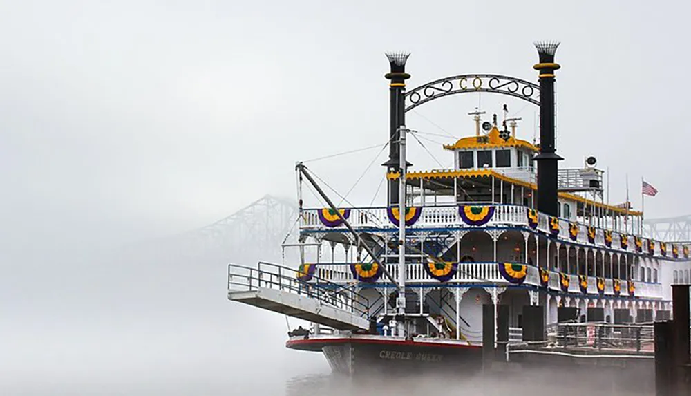 A paddlewheel riverboat is moored at a foggy riverside with a barely visible bridge in the background