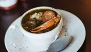 The image shows a savory seafood soup with a prominent crab leg and other ingredients served in a white bowl with a spoon on the side.