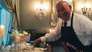 A smiling person is pouring wine into glasses at an elegantly set table with candles and a bucket of chilled bottles in the background.