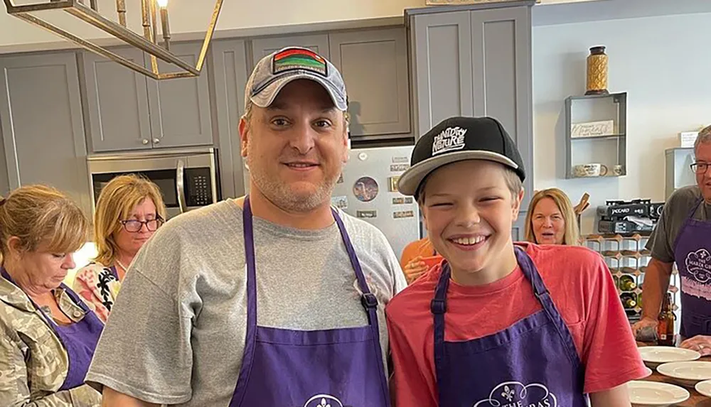 A man and a young boy wearing matching purple aprons and smiling are posing for a photo in a kitchen setting with other people around them possibly during a cooking class or family event