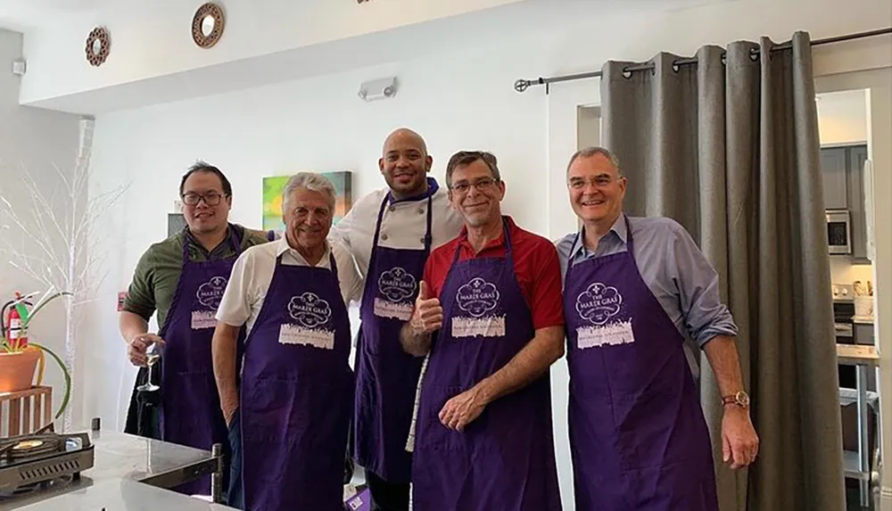 Five smiling people wearing matching purple aprons pose together in what appears to be a kitchen setting