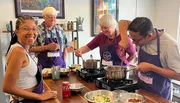 Four people are happily engaged in a cooking class, wearing purple aprons and preparing food on a kitchen countertop.