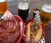 The image depicts a laid-back meal setting with a large soft pretzel a sandwich loaded with toppings and three glasses of different types of beer on a wooden table