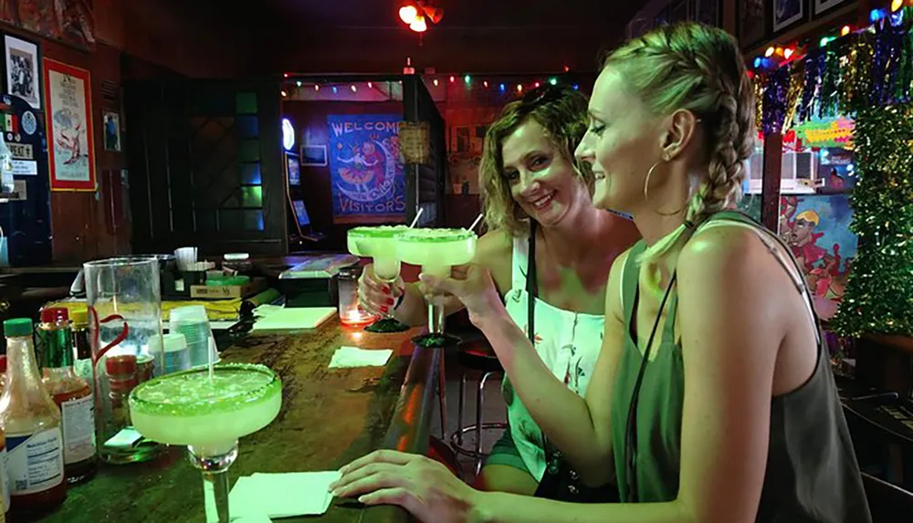 Two people are cheerfully toasting with large margarita glasses in a festive bar setting