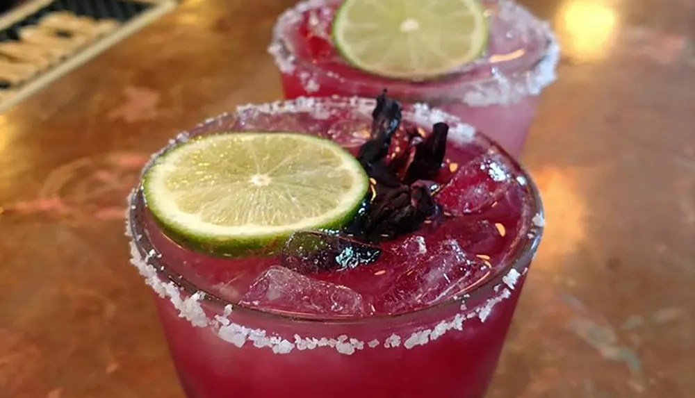 The image shows two glasses of a reddish-purple cocktail with salt rims and lime garnishes on top of a bar counter