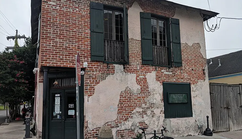 An old brick building with weathered green shutters and patches of missing plaster showing a glimpse of the very storied and textured history on its facade