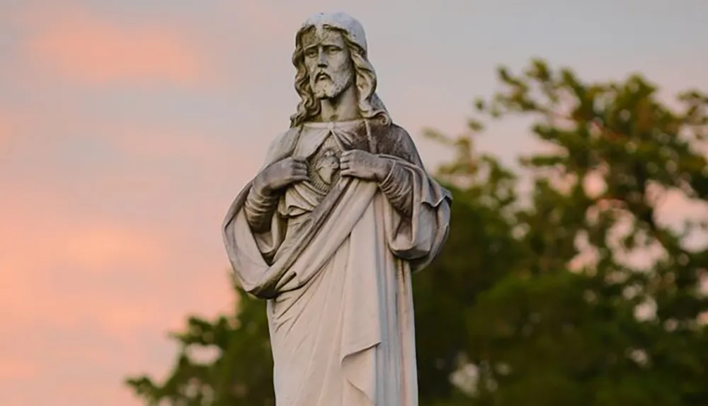 The image shows a statue of a bearded figure with long hair traditionally dressed hands over chest with a heart visible in the center set against a dusky sky and foliage in the background