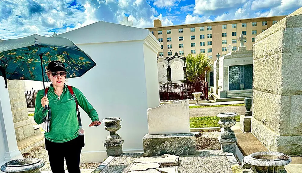 A person holding an umbrella stands among ornate gravesites on a sunny day with a blue sky overhead