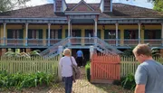 Two individuals approach the gated entrance of a colorful historic two-story house with decorative trim and a wraparound porch.