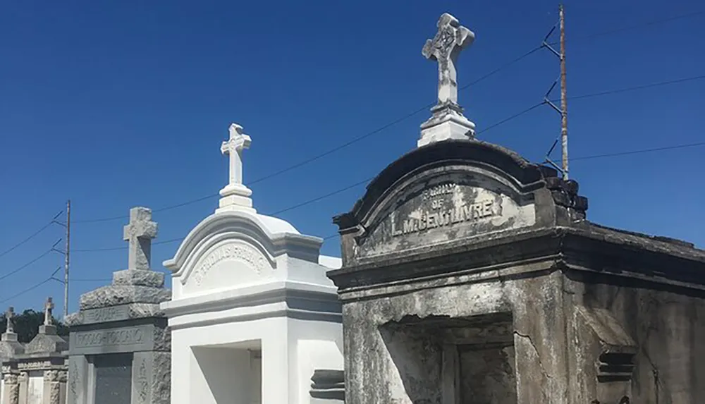 The image displays a series of ornate above-ground tombs with crosses typical of a historic cemetery under a clear blue sky