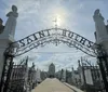 The image shows an ornate entrance gate to Saint Roch Campo Santo cemetery with statues on either side and a sunlit path leading into the burial grounds