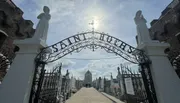 The image shows an ornate entrance gate to Saint Roch Campo Santo cemetery with statues on either side and a sunlit path leading into the burial grounds.