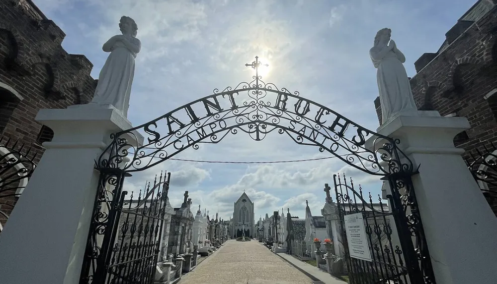 The image shows an ornate entrance gate to Saint Roch Campo Santo cemetery with statues on either side and a sunlit path leading into the burial grounds