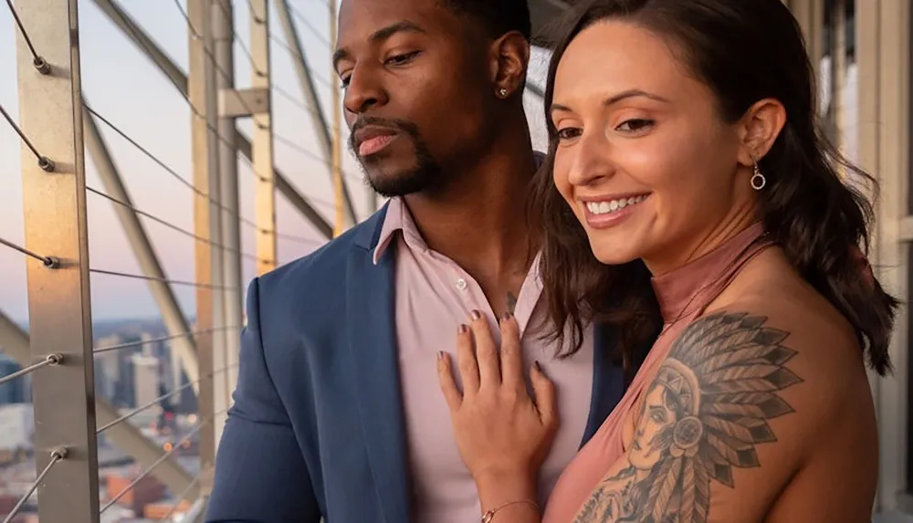 A man and a woman are sharing a tender moment together with the woman smiling and resting her hand on the mans chest showcasing her prominent arm tattoo as they both gaze out of a balcony with a cityscape in the background