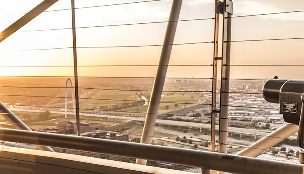 The image showcases a sunset view from an elevated observation point featuring cable barriers a telescope and a distant arch structure in the landscape