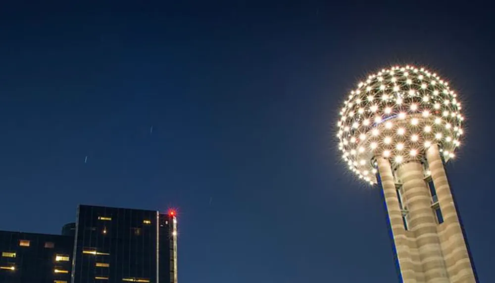 The image captures a distinctive illuminated spherical structure atop a columnar base against a night sky alongside a modern building with lit windows