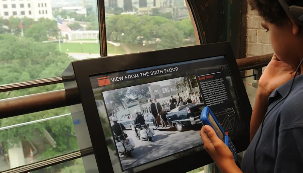 A person is using an interactive display titled VIEW FROM THE SIXTH FLOOR with historical content while standing by a window overlooking a park