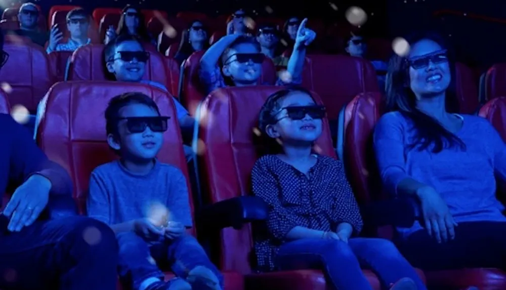 A group of people including children and adults are wearing 3D glasses and enjoying a movie in a theater with red seats