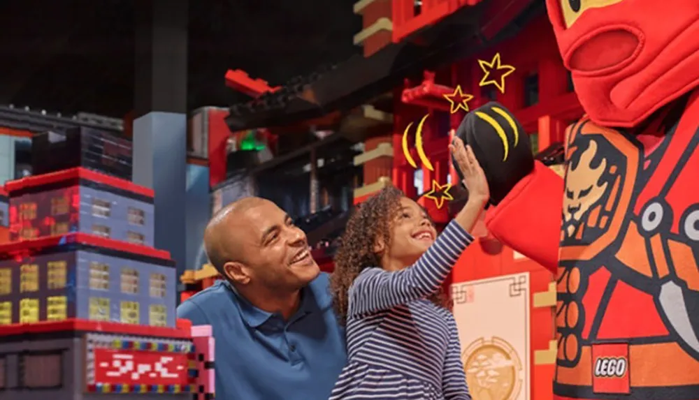 A man and a young girl are smiling and high-fiving a life-size LEGO character in a vibrant LEGO-themed environment