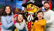 A family is posing with a life-sized Lego pirate figure, all smiling cheerfully in a colorful, playful setting.