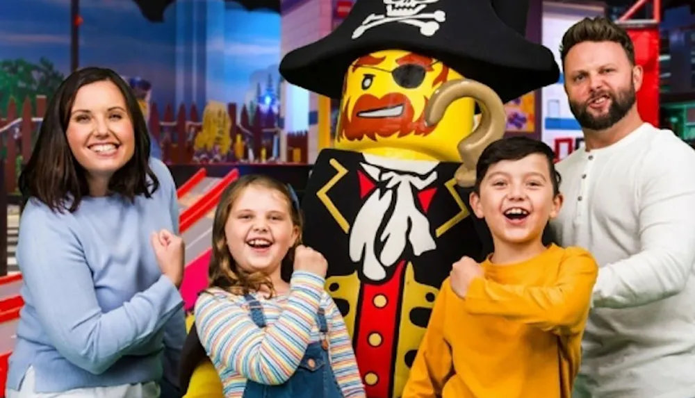 A family is posing with a life-sized Lego pirate figure all smiling cheerfully in a colorful playful setting