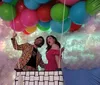 Two people pretend to float amidst colorful balloons against a whimsical backdrop of clouds and a spectrum of pastel colors