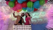 Two people pretend to float amidst colorful balloons against a whimsical backdrop of clouds and a spectrum of pastel colors.