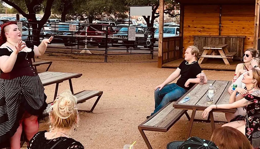 An animated individual is performing or speaking to a casual outdoor audience seated at picnic tables