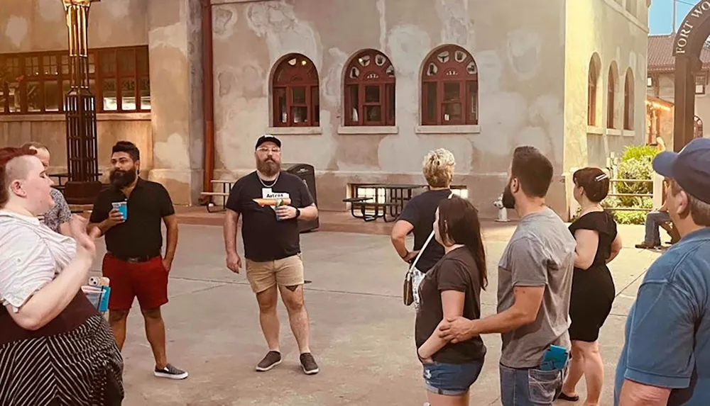 A group of people appears to be engaged in conversation and enjoying beverages outdoors in a public space with European-style buildings in the background