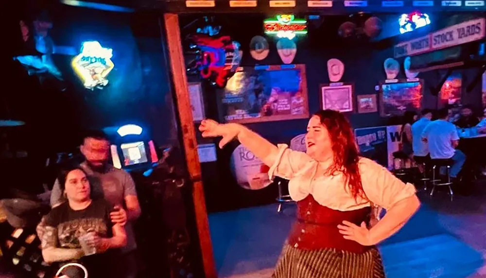 A person in a traditional outfit is animatedly singing or speaking with one arm extended with onlookers in a dimly-lit bar-like setting adorned with neon signs and posters