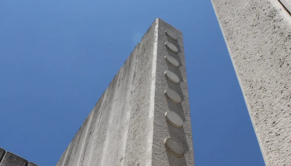 The image captures a low-angle view of a modern concrete structure with rows of circular embellishments set against a clear blue sky