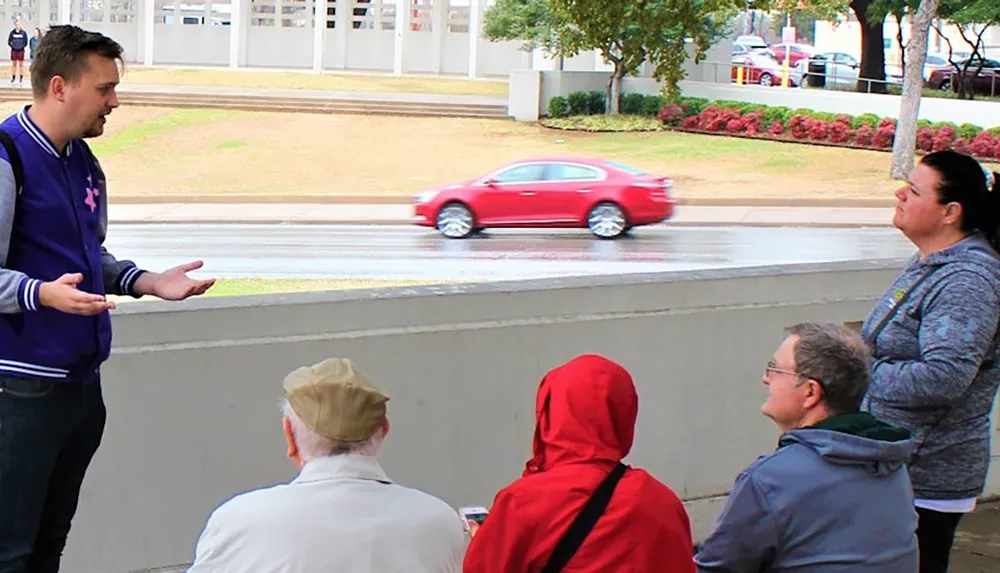 A group of people including a gesturing man appears to be engaged in an outdoor conversation by a street with passing traffic