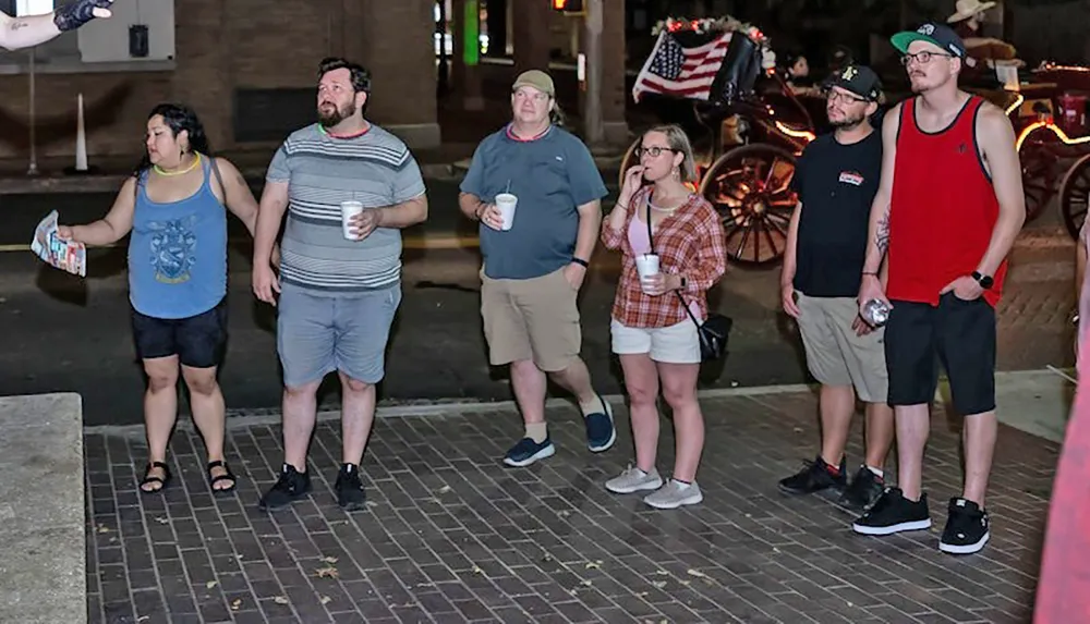 A group of people is standing idly on a street at night with some holding drinks and a horse-drawn carriage adorned with an American flag visible in the background