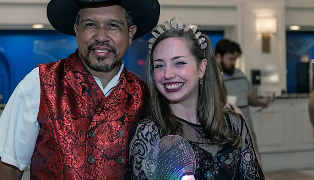 Two people are smiling at a social event one wearing a cowboy hat and a festive red vest and the other with a lace dress and a crown-like headband holding a light-up orb