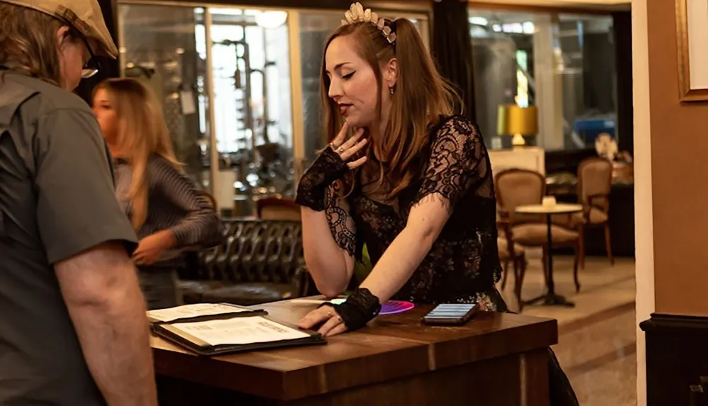 A woman dressed in a lace dress is standing behind a podium engaging in conversation with a person partially visible on the left side of the frame in what appears to be a warmly lit reception or lounge area